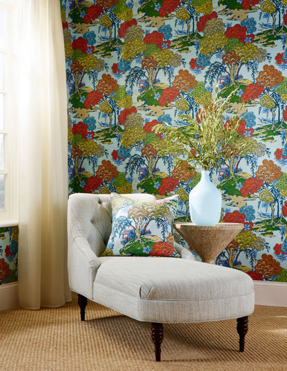 Cream chaise lounge with dark legs, round wood table and white vase; bold multicolor tree wallpaper, pillows, jute rug.