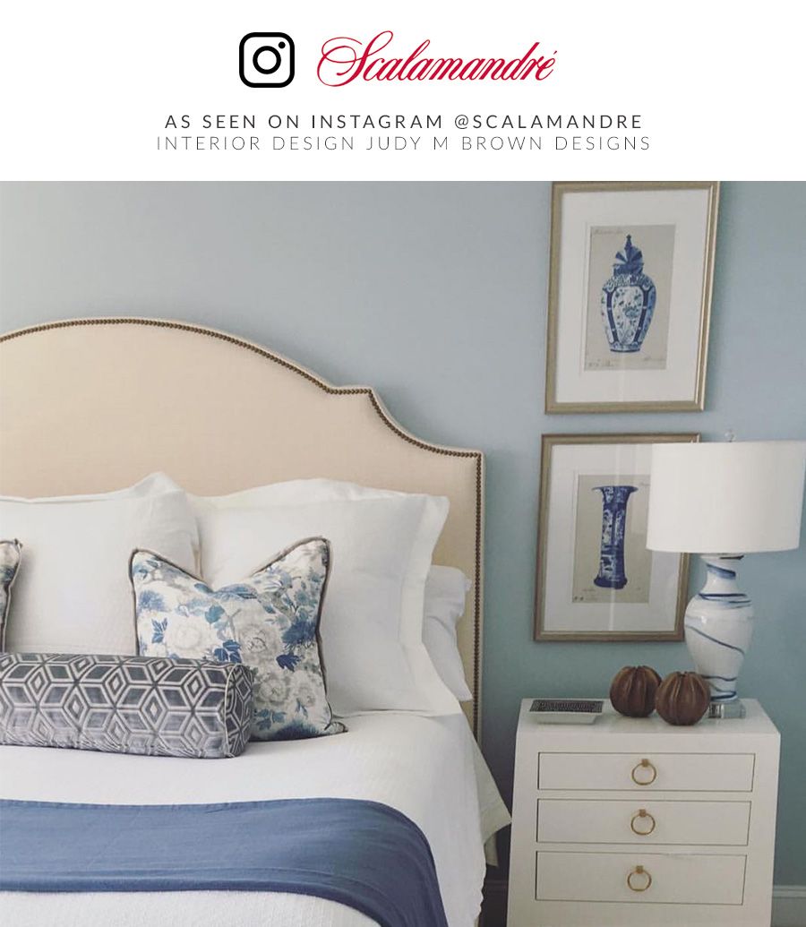 Coastal bedroom with beige upholstered headboard, white linens, blue throw, mixed pillows, white nightstand, blue lamp.