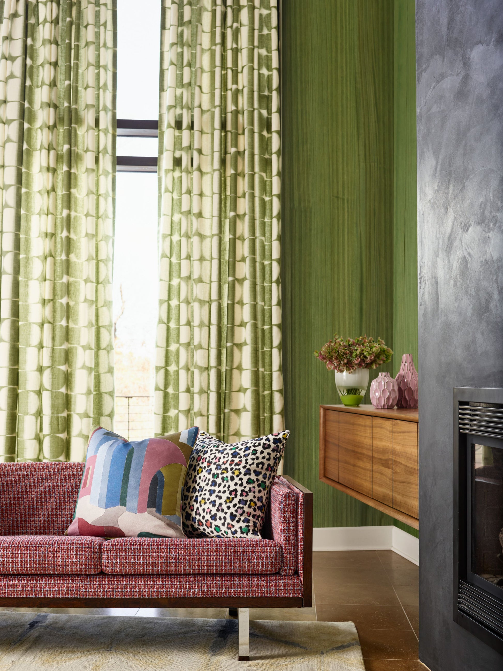 Red textured sofa with colorful geometric and animal-print pillows, green curtains, green wall, console with pink vases.