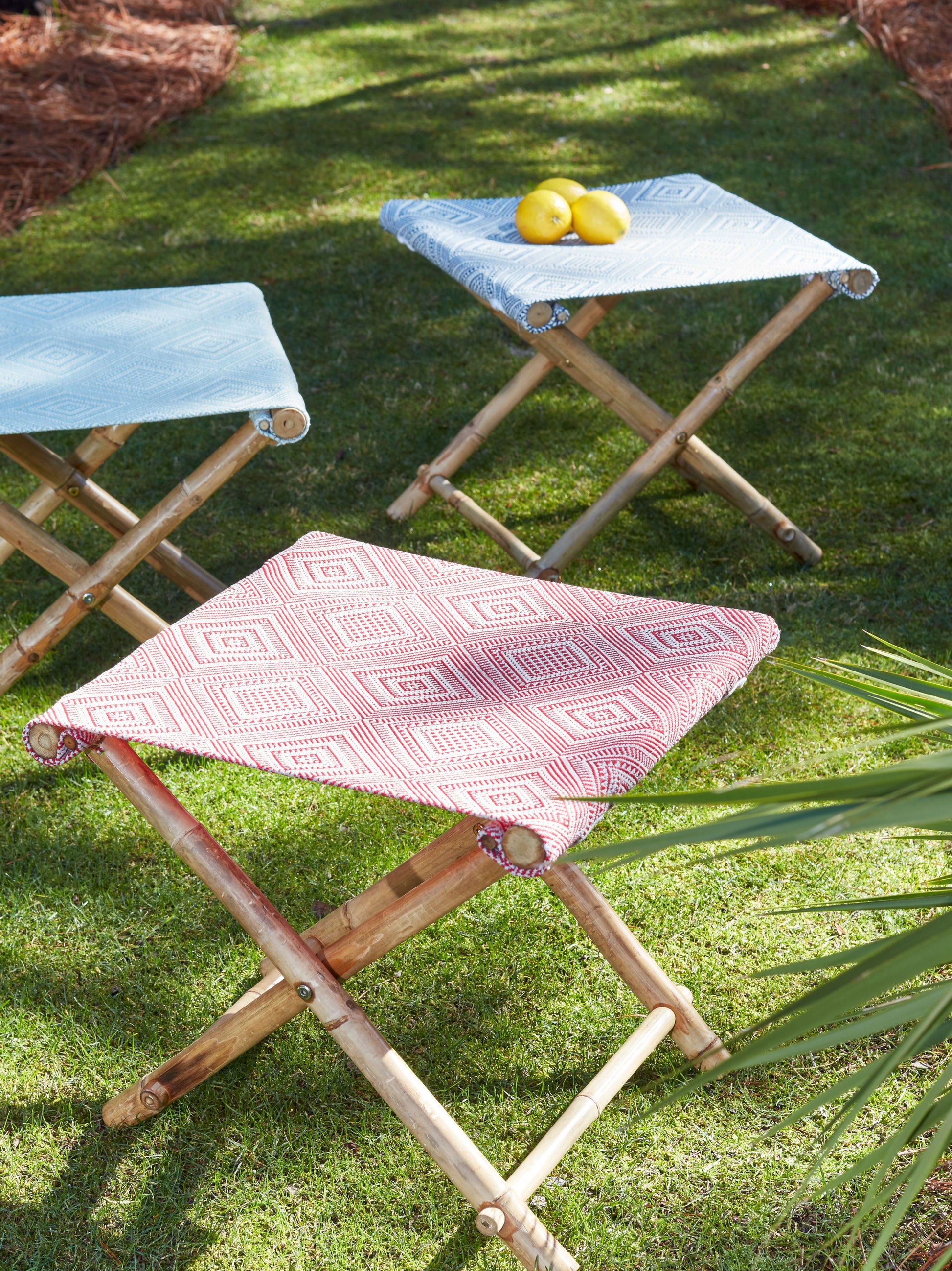 Three bamboo folding stools with blue and red diamond-patterned fabric seats; lemons on one in a sunny garden.