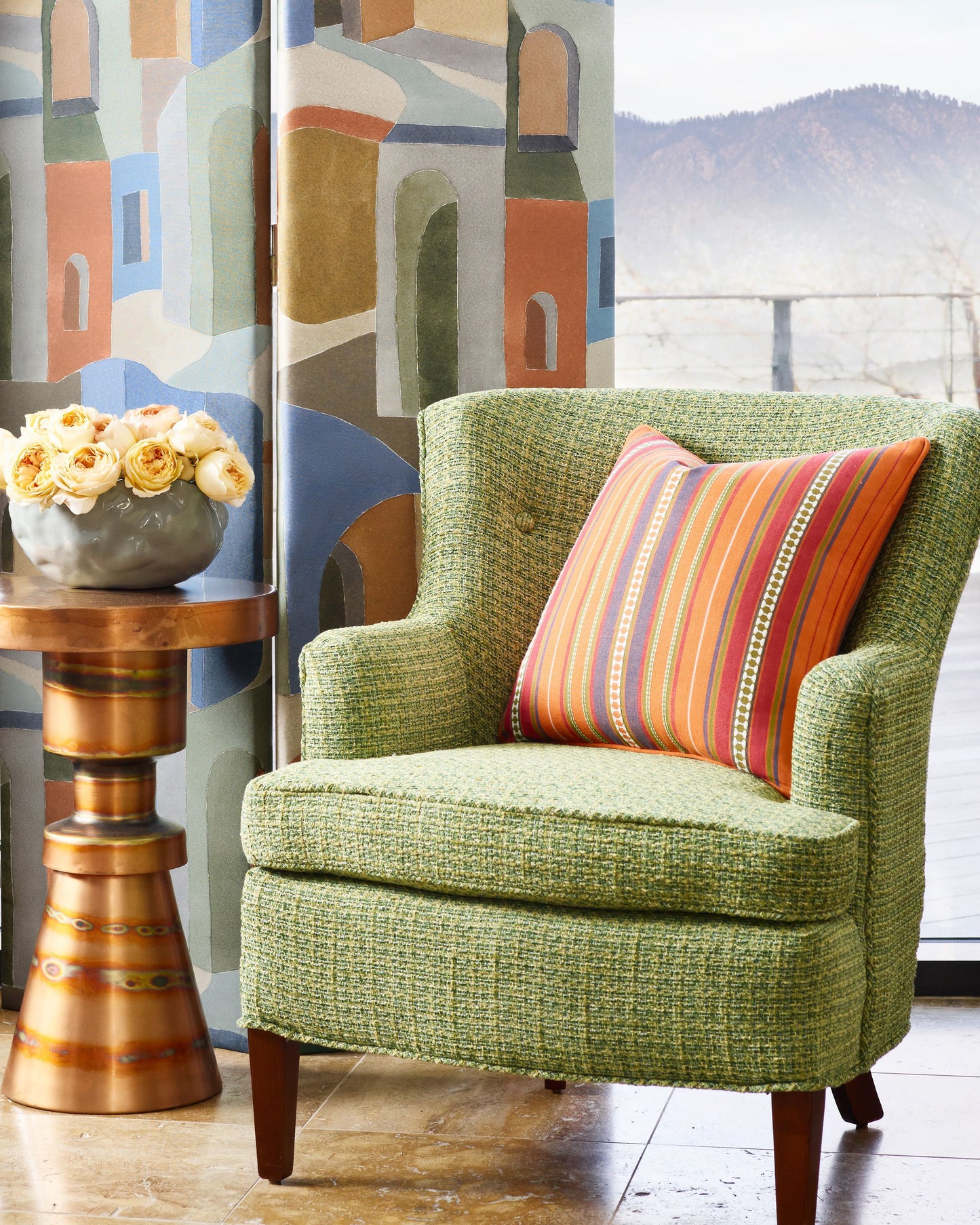 Green tweed armchair with striped orange pillow, beside a wooden pedestal table with pale yellow roses, mural backdrop.