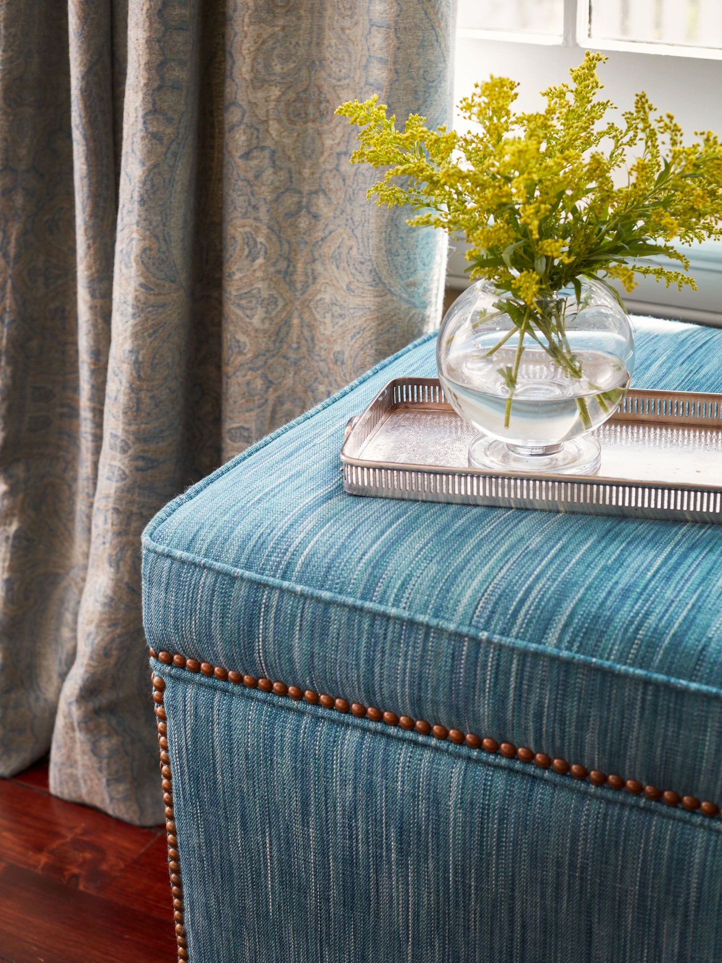 Turquoise upholstered ottoman with nailhead trim, topped by a silver tray and glass vase of yellow flowers.