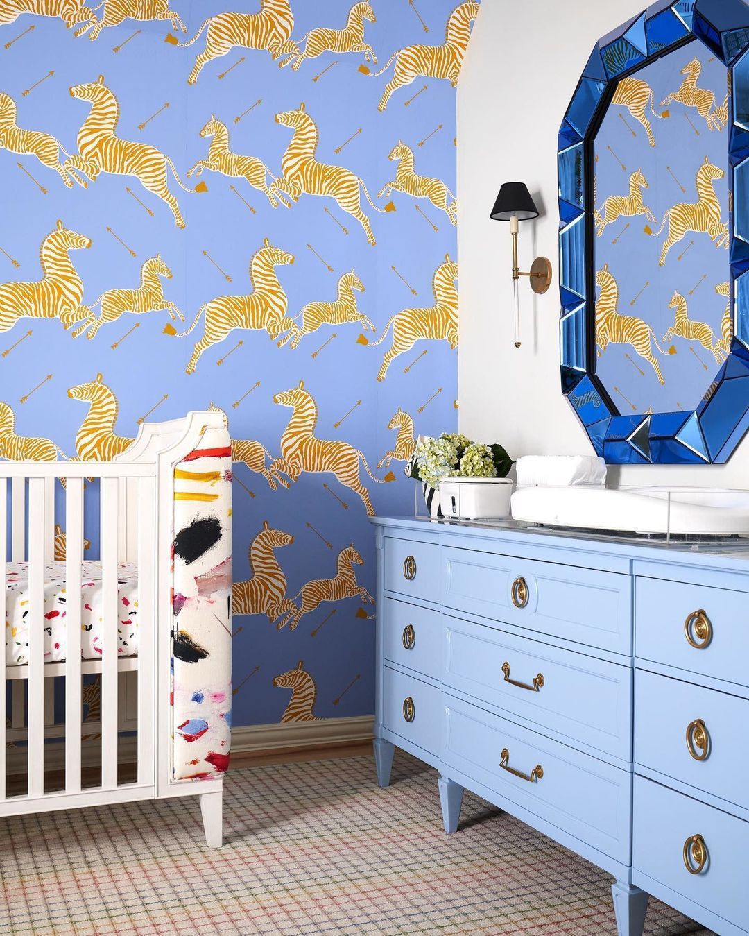 Nursery with periwinkle zebra wallpaper, light-blue dresser, white crib, and blue geometric mirror.
