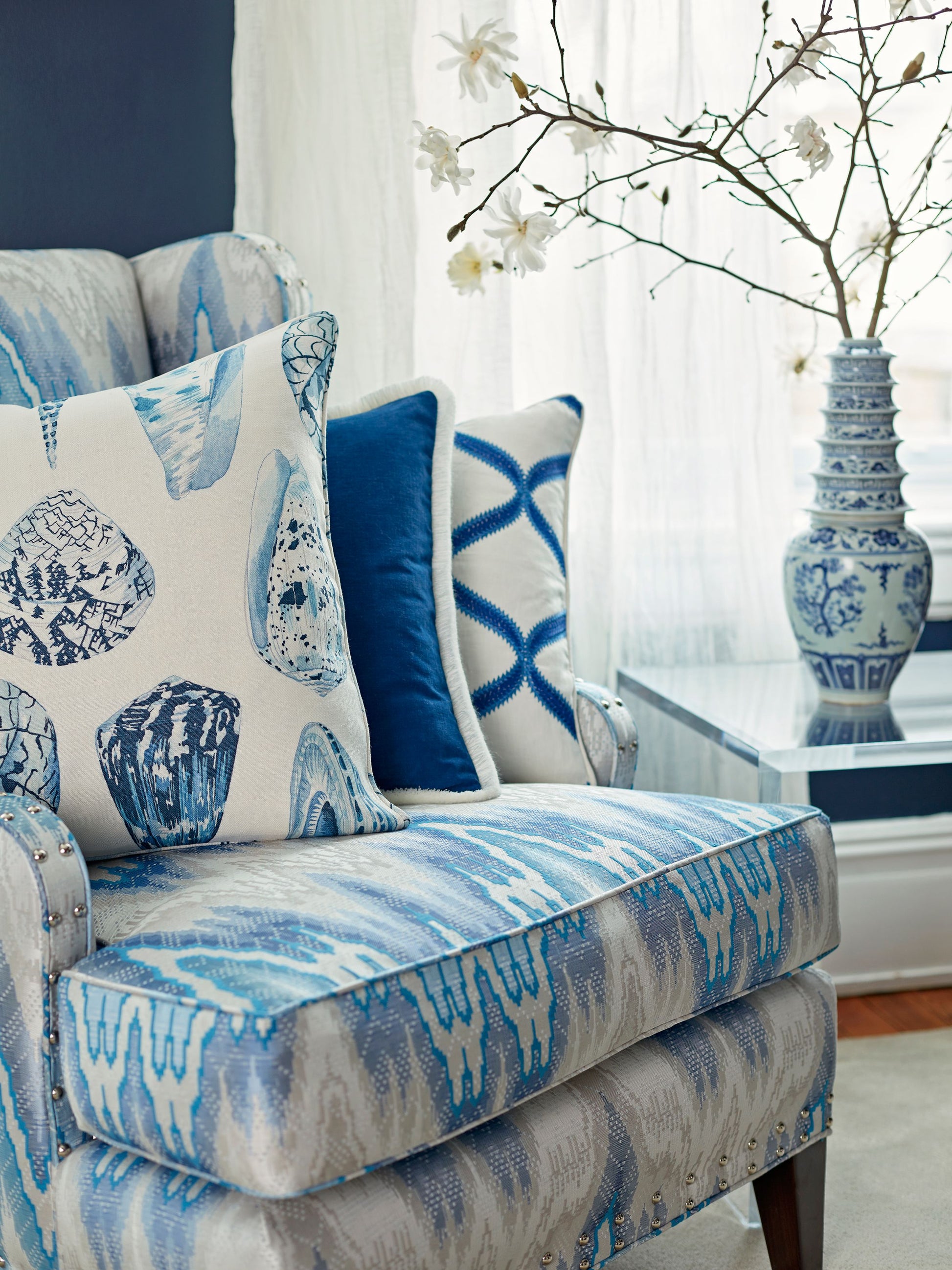 Blue-and-white coastal chair with ikat upholstery and shell-print cushions, glass table with blue vase and blossoms.