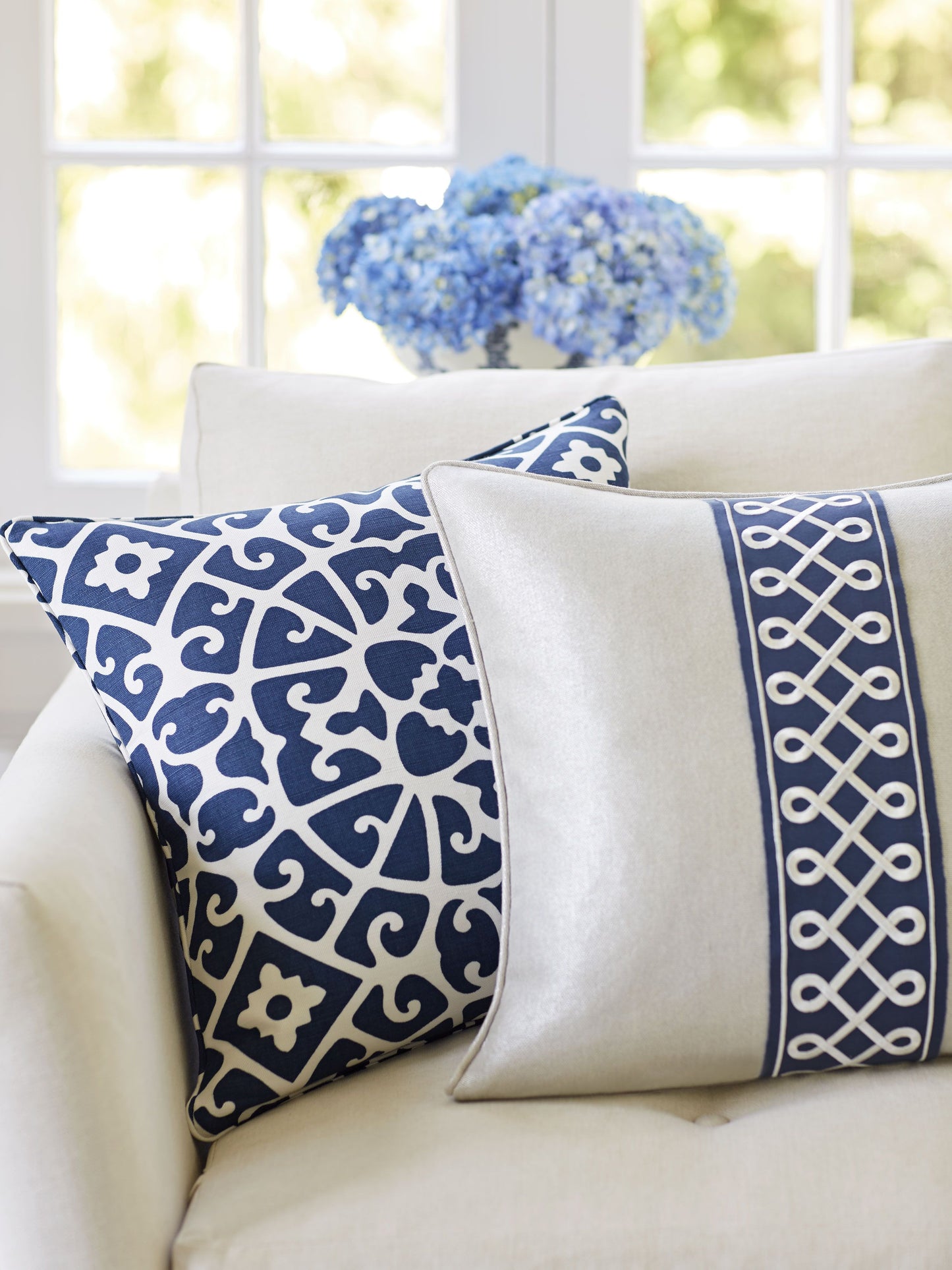 Two decorative throw pillows on a cream sofa: navy scroll pattern and cream with navy lattice trim, blue hydrangeas in back.