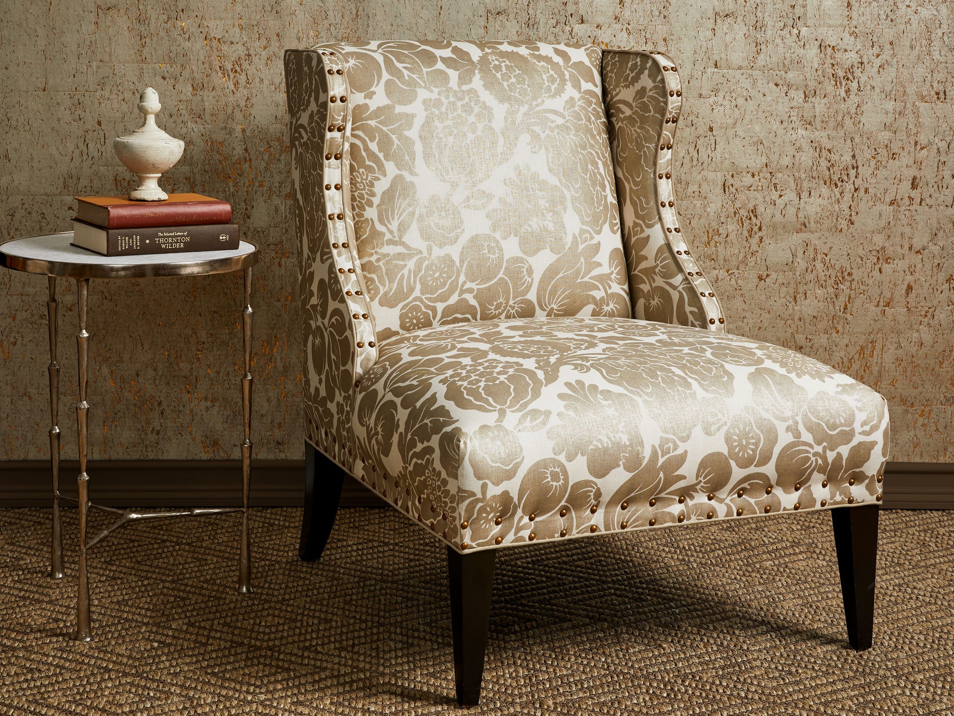 Beige damask upholstered wingback chair with nailhead trim and dark tapered legs beside a round marble-top side table with books and a white vase.