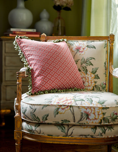 Pink diamond-pattern throw pillow with green pom-pom trim on a vintage floral upholstered wooden armchair.