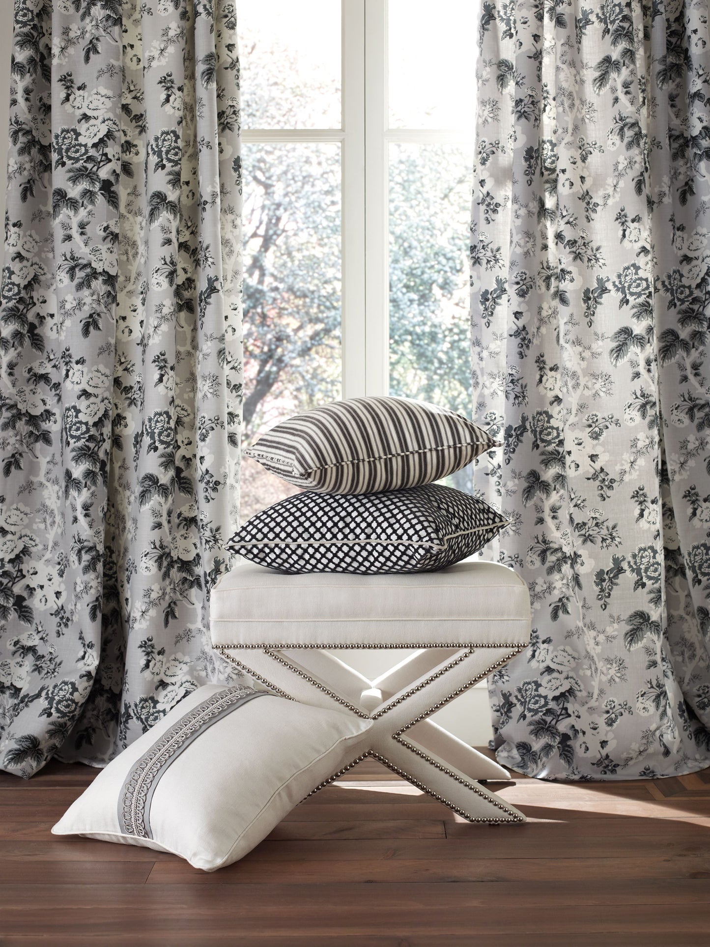 Stack of striped, dotted, and geometric cushions on a cream tufted ottoman, with gray floral curtains and wooden floor.