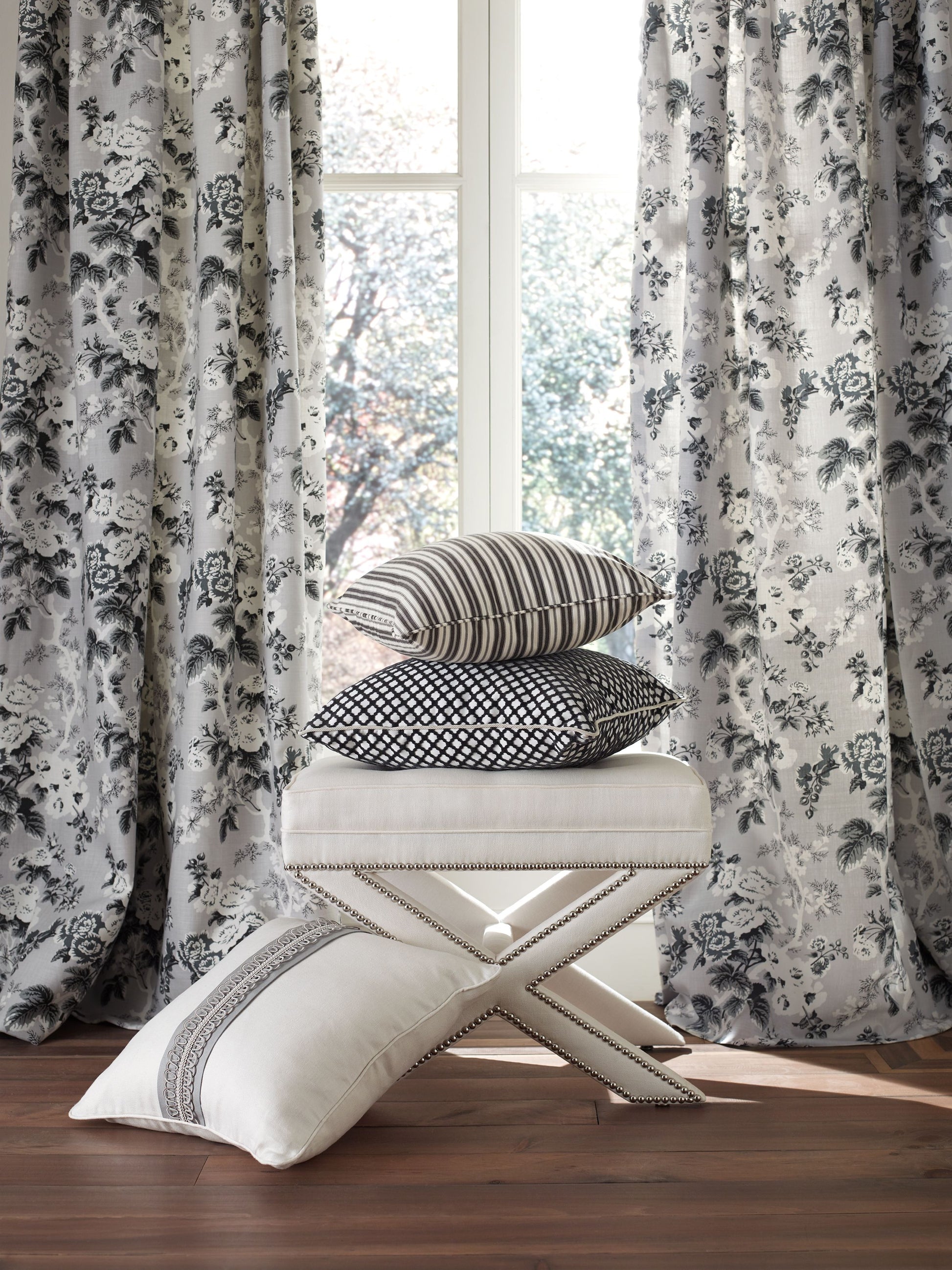 Stack of striped, dotted, and geometric cushions on a cream tufted ottoman, with gray floral curtains and wooden floor.