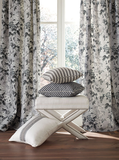 Stack of striped, dotted, and geometric cushions on a cream tufted ottoman, with gray floral curtains and wooden floor.