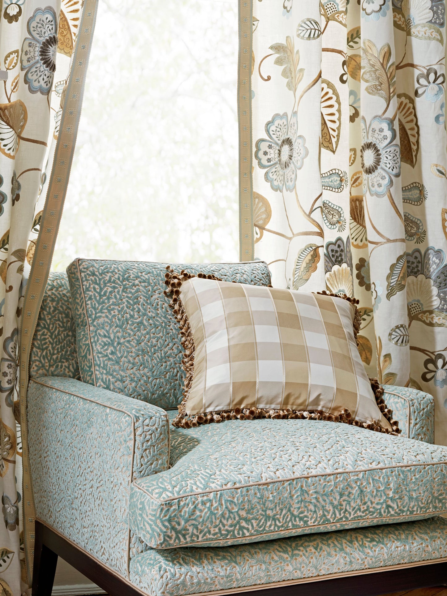 Teal patterned armchair with beige gingham cushion edged in brown fringe, beside cream floral curtains.