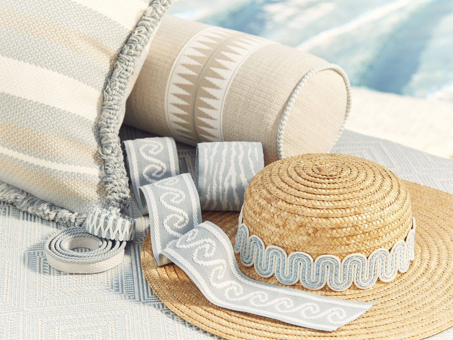 Straw sun hat with light blue wavy trim beside striped cushions and rolled ribbons on a woven outdoor rug.