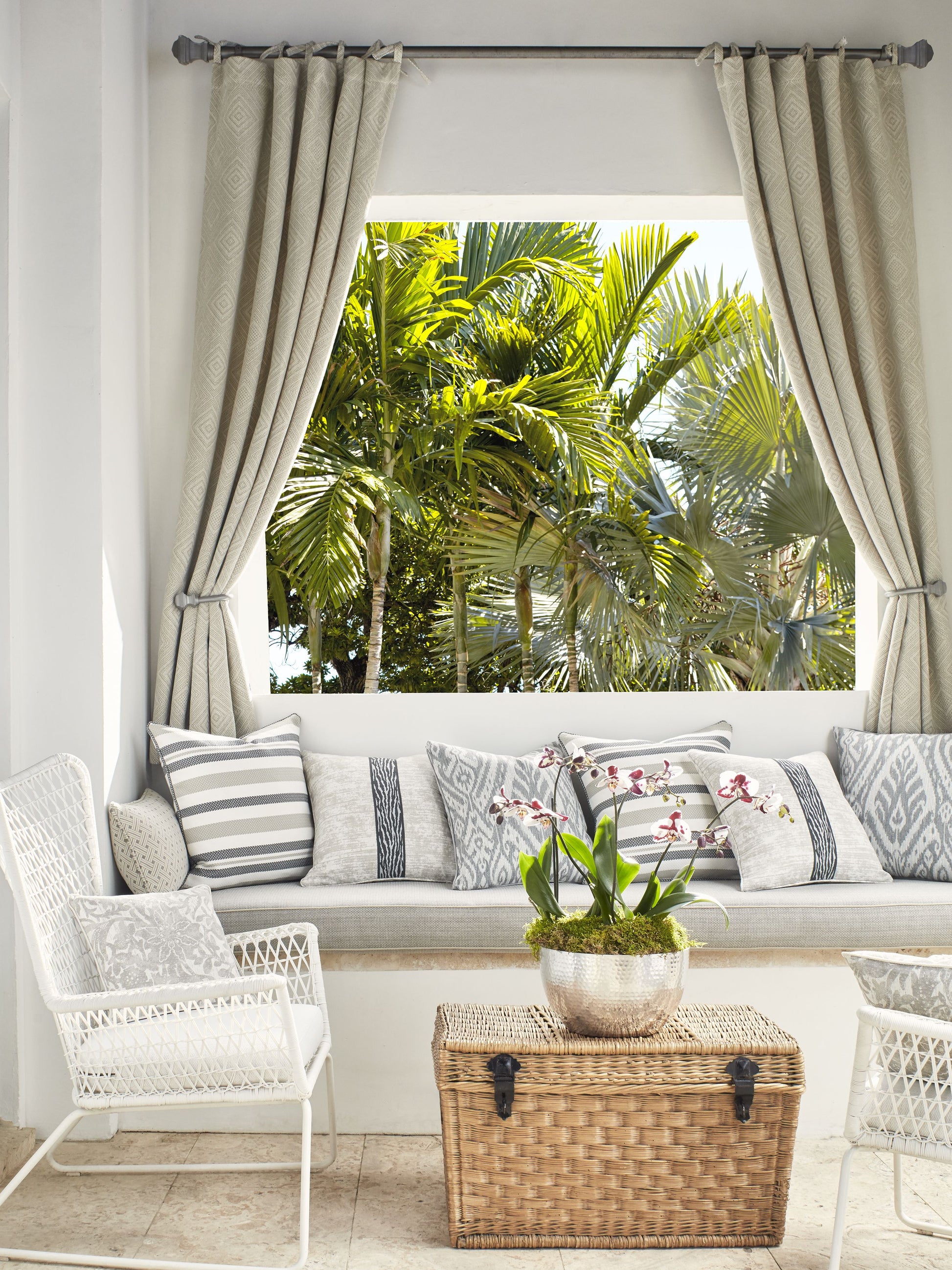 Neutral outdoor lounge with white wicker chairs, cushioned bench, striped pillows, wicker trunk table, orchid plant, palm view