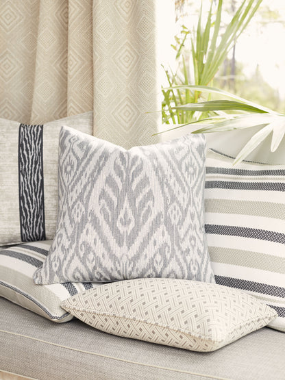 Gray ikat-patterned throw pillow atop a neutral striped sofa with woven cushions and beige diamond curtains.