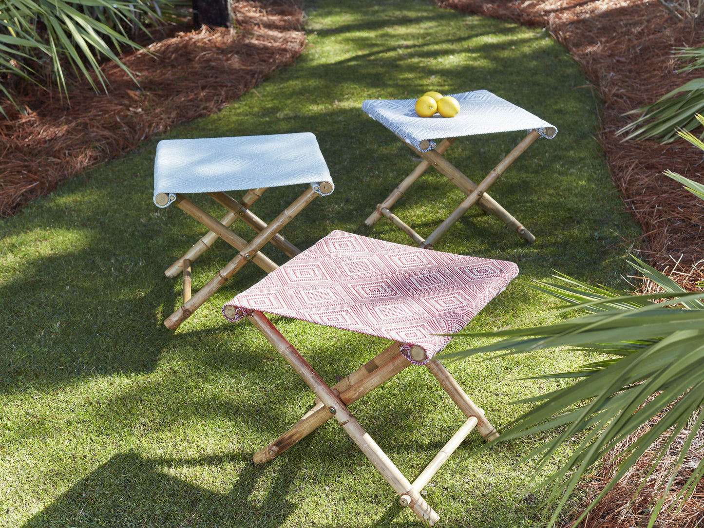 Three bamboo folding stools with blue and red woven seats on a grassy garden lawn; one top with lemons.