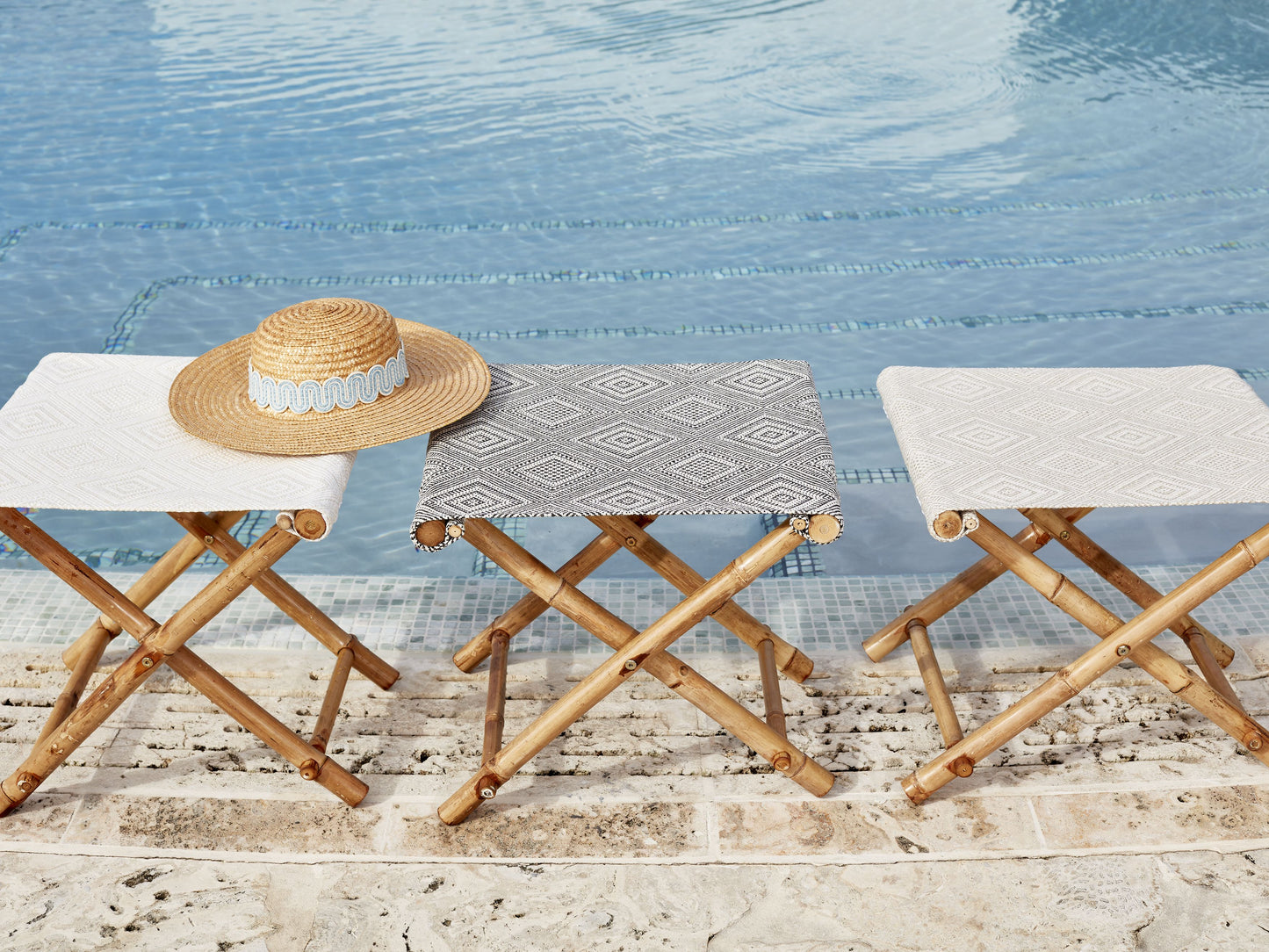 Three wooden folding stools with white and gray patterned seats by a pool; straw hat rests on the left stool.