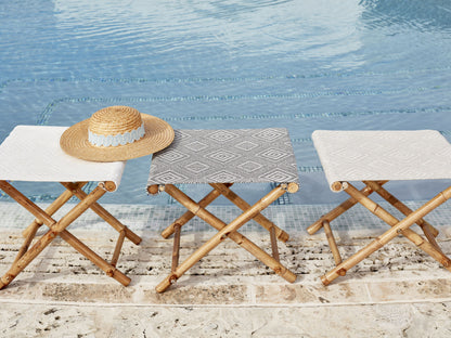 Three wooden folding stools with white and gray patterned seats by a pool; straw hat rests on the left stool.