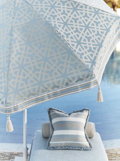 Blue geometric-pattern patio umbrella with white lattice and tassel trim; striped poolside cushions on a lounge.