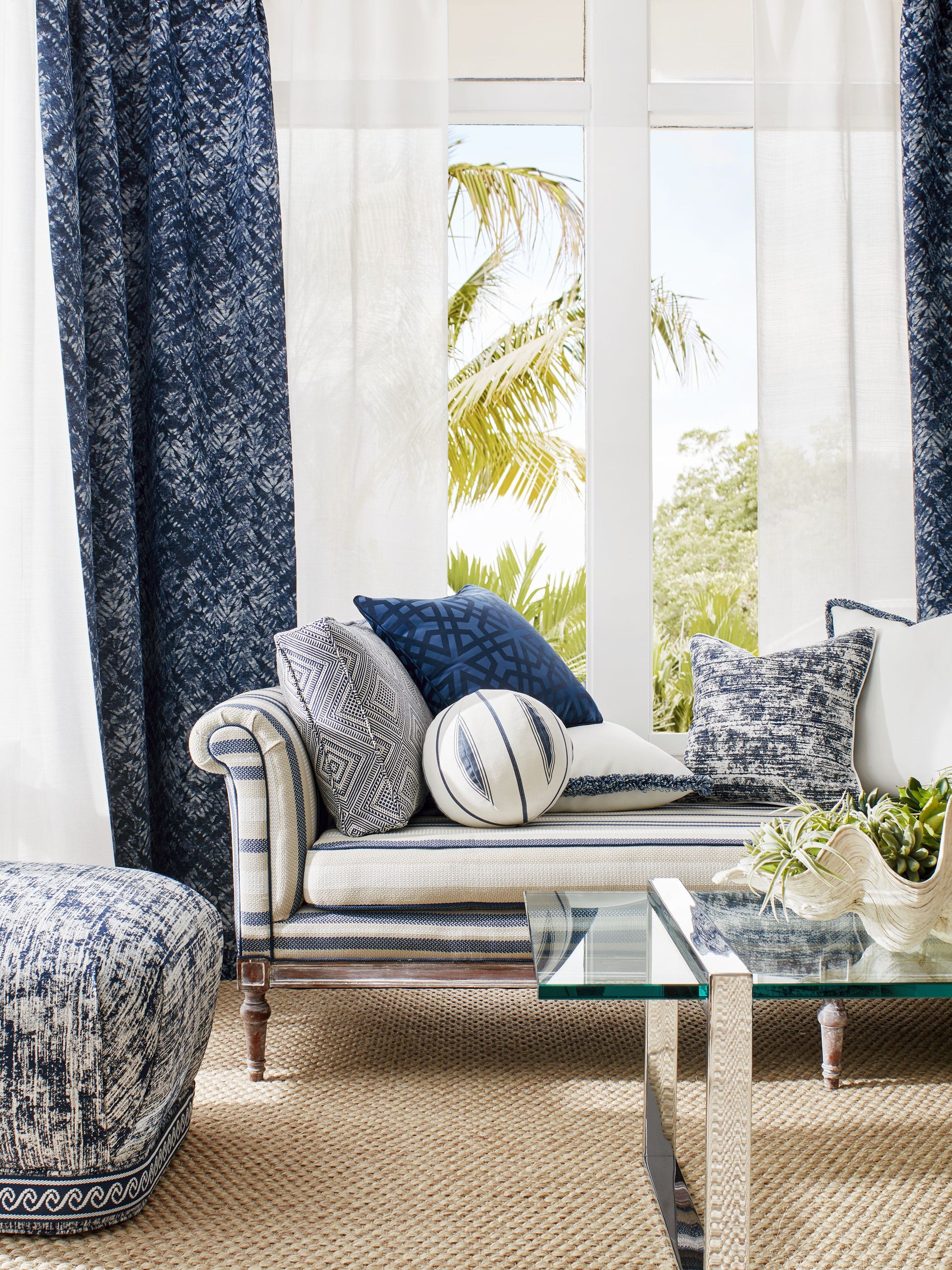 Coastal living room: navy and cream striped sofa, blue cushions, sheer white and navy curtains, glass coffee table, jute rug.