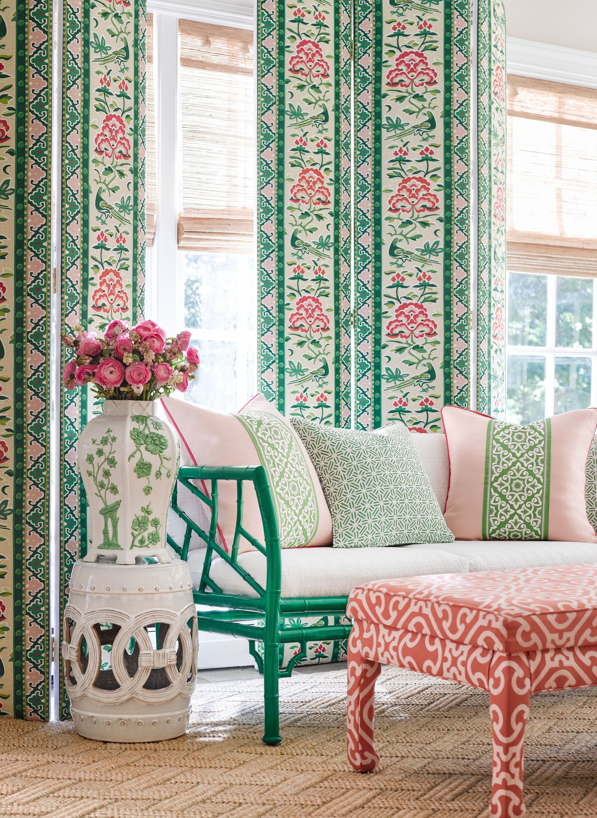 Teal bamboo chair with pink cushions beside floral striped wallpaper, white vase with pink flowers and coral ottoman on jute