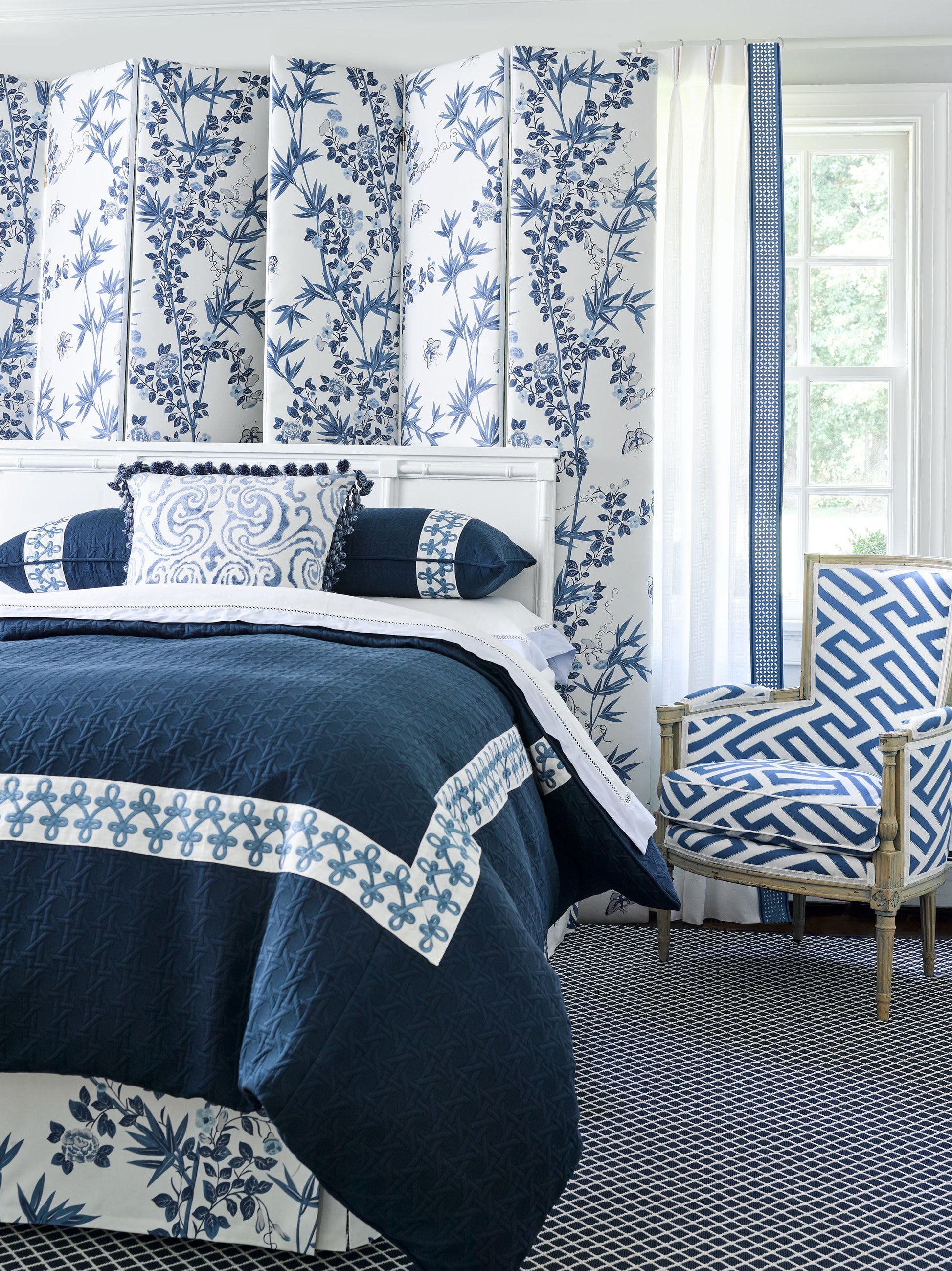 Navy quilt with white border and blue lattice trim on white bed, blue botanical wallpaper, and blue geometric chair.