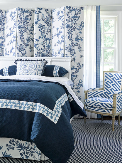 Navy quilt with white border and blue lattice trim on white bed, blue botanical wallpaper, and blue geometric chair.