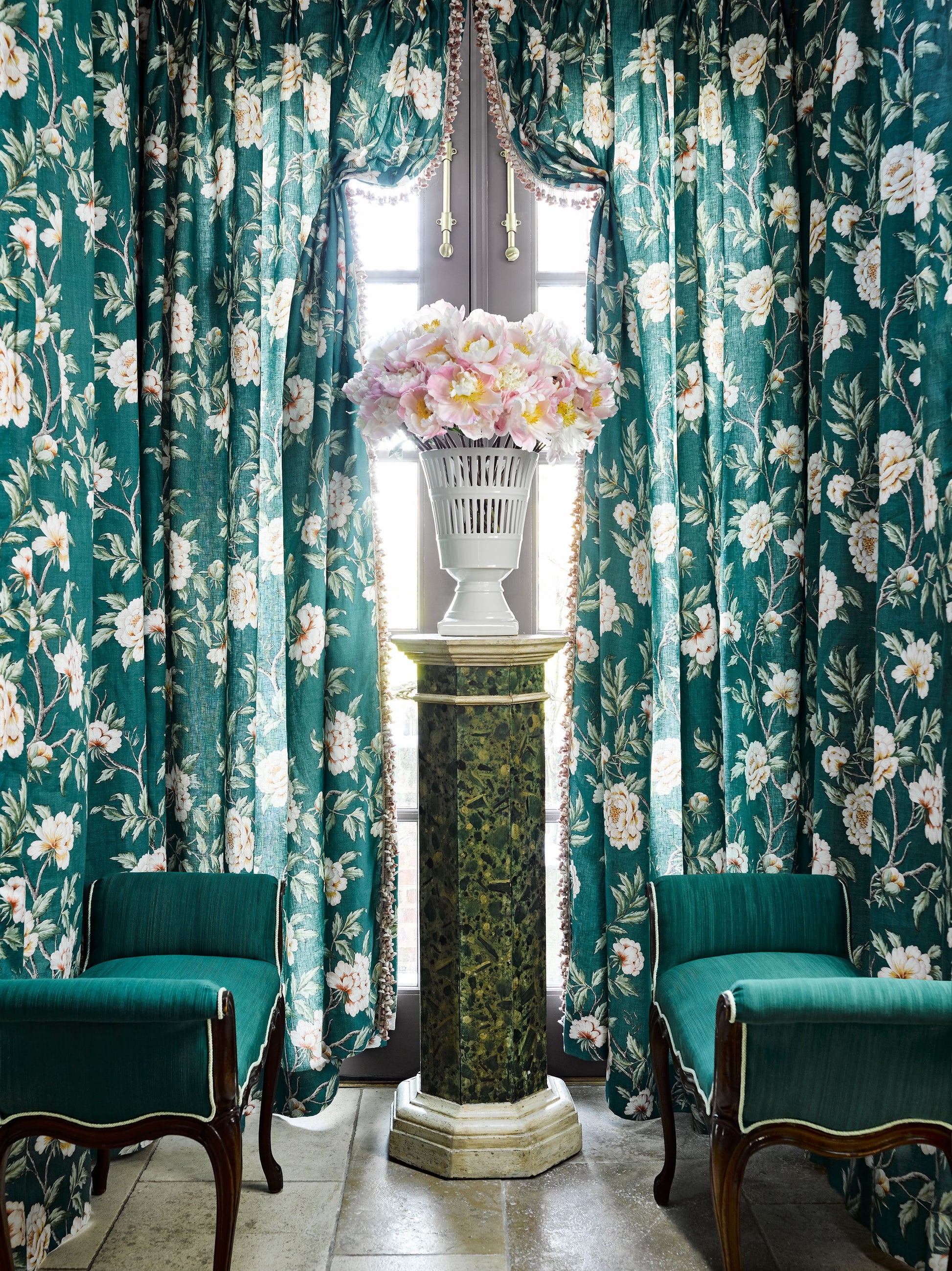 Teal floral curtains frame a marble pedestal with a white vase of pink & white hydrangea, flanked by green velvet armchairs