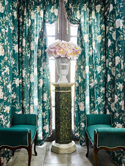 Teal floral curtains frame a marble pedestal with a white vase of pink & white hydrangea, flanked by green velvet armchairs