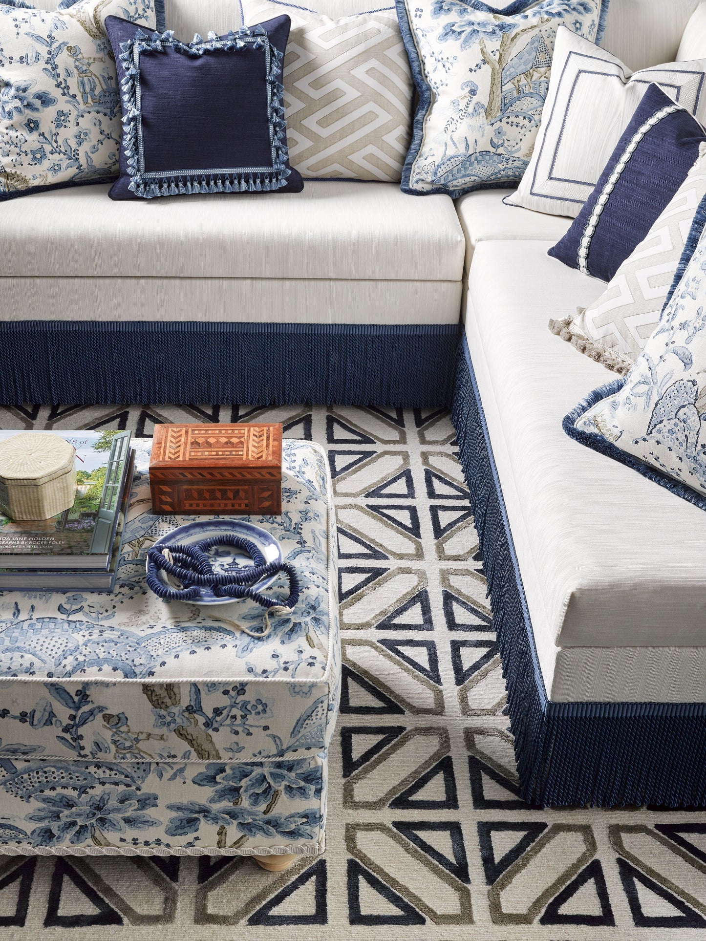 Navy & white sectional with floral & geometric cushions, blue fringe trim, geometric rug; ottoman with books & wooden box.