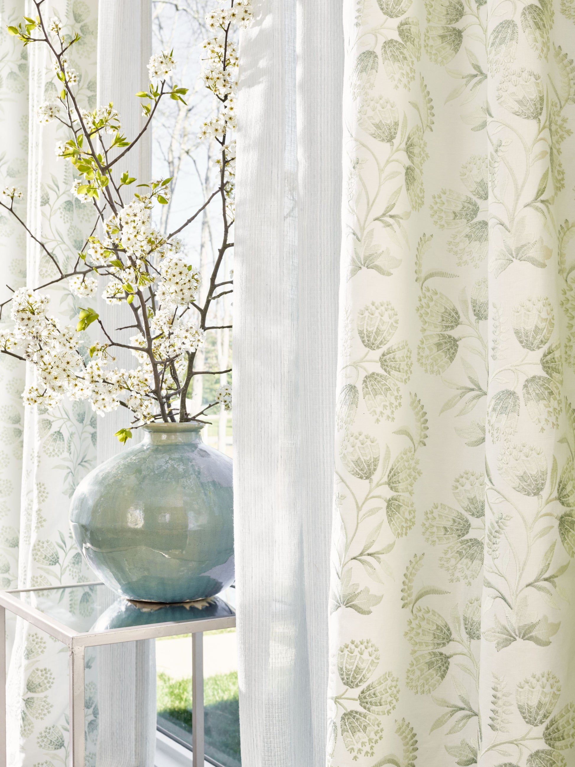 Soft sheer curtains frame pale green botanical drapes; blue glazed ceramic vase with flowering branches on a glass console.