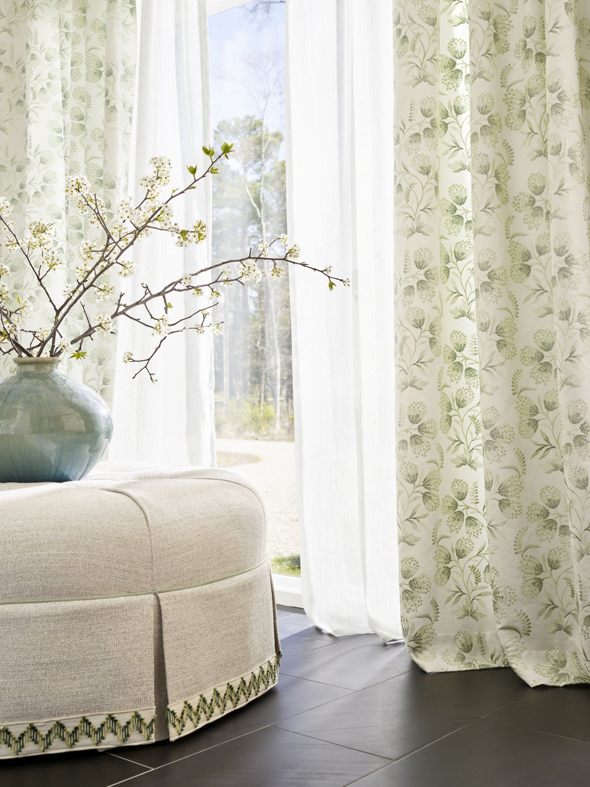 Sheer white and botanical curtains as the focal point, with a beige upholstered ottoman with green trim and a blue ceramic vase of white blossoms beside.