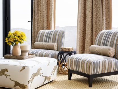 Beige-blue striped upholstered lounge chairs with bolster pillows, leopard-print ottoman, wicker side table, yellow bouquet.