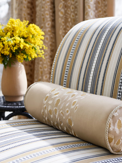 Beige striped armchair with gold-embroidered cylindrical bolster; yellow bouquet in a vase behind.