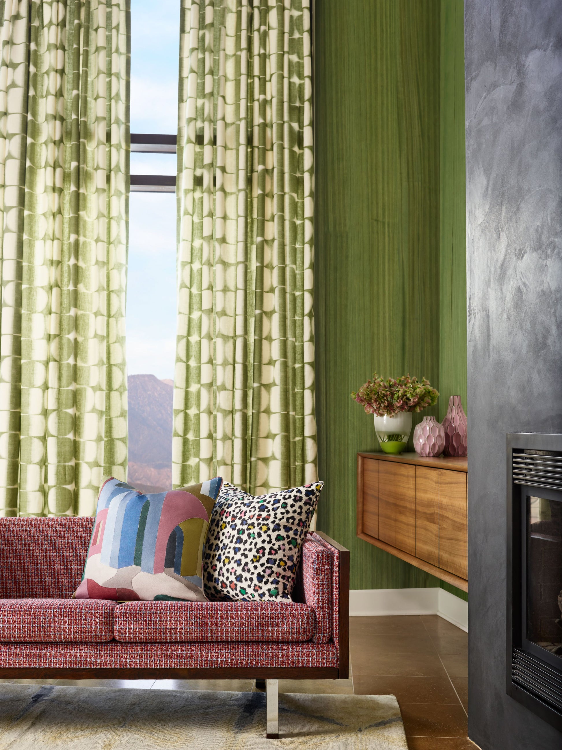 Red textured sofa with colorful geometric and leopard print pillows, green patterned curtains, wooden sideboard, and a fireplace.