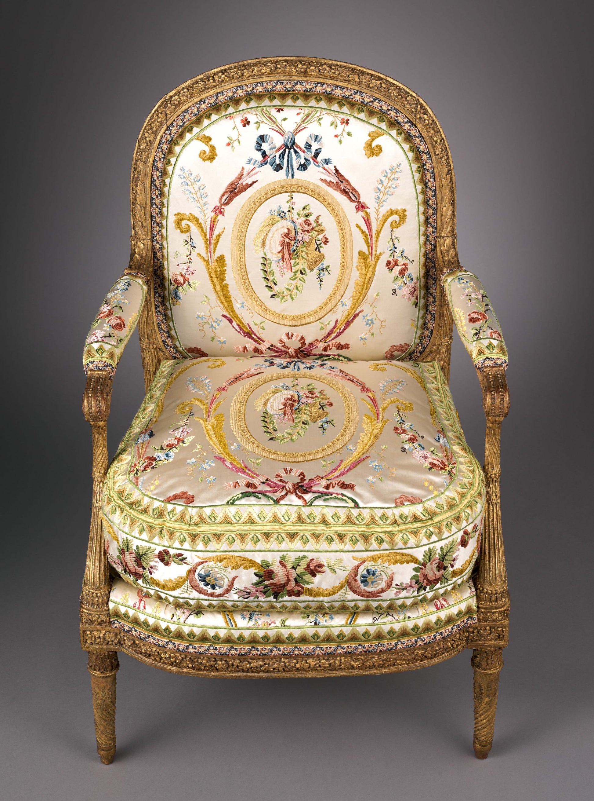 Ornate gilded wood armchair with carved frame and cream silk upholstery featuring floral medallions and scrollwork.