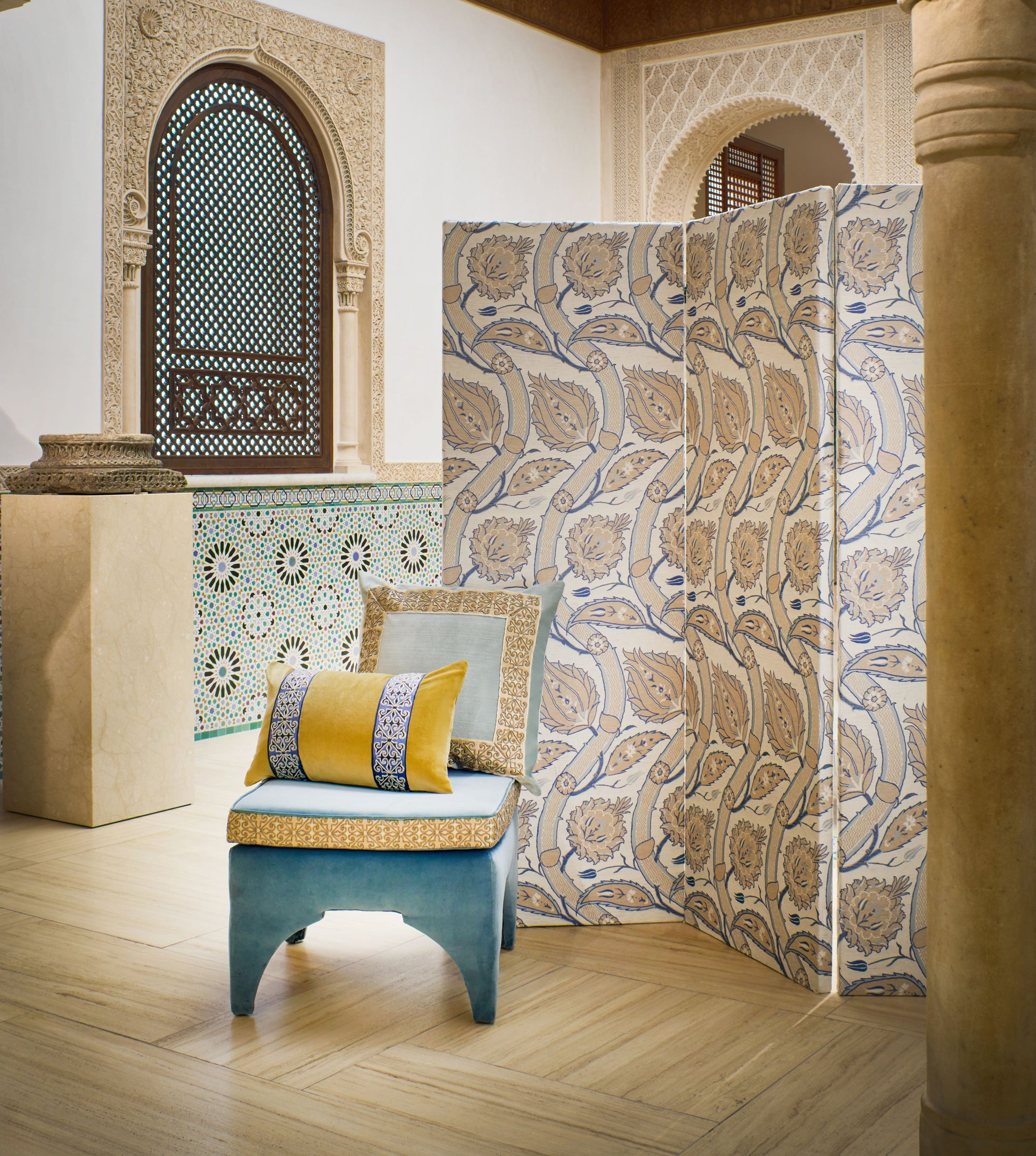 Three-panel blue-and-gold floral folding screen beside a pale blue ottoman with yellow cushions in a Moroccan-inspired interior.