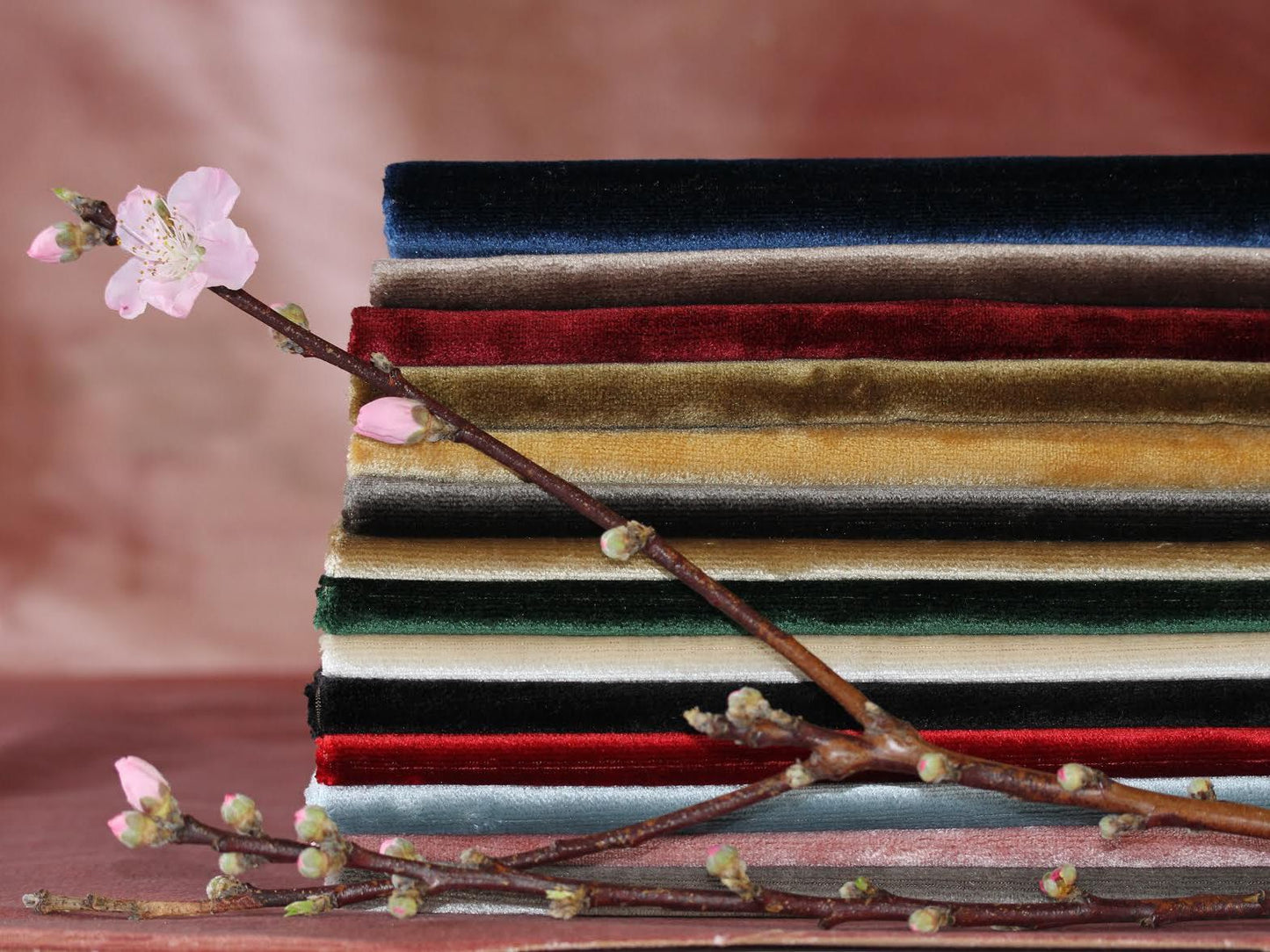 Stack of velvet fabrics in navy, red, gold, olive, cream, and black with a pink blossom branch laid across.