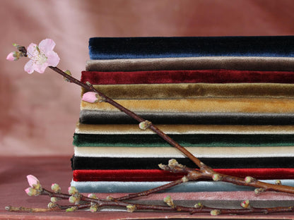 Stack of velvet fabrics in navy, red, gold, olive, cream, and black with a pink blossom branch laid across.