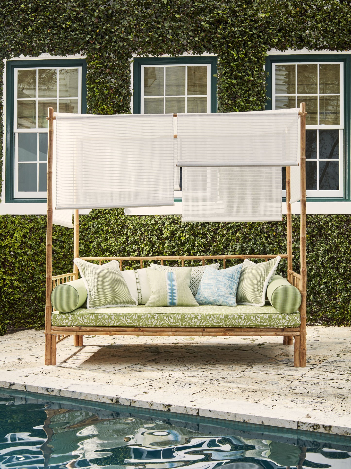Wood outdoor daybed with white fabric screens, green patterned cushions, assorted pillows, poolside under ivy.