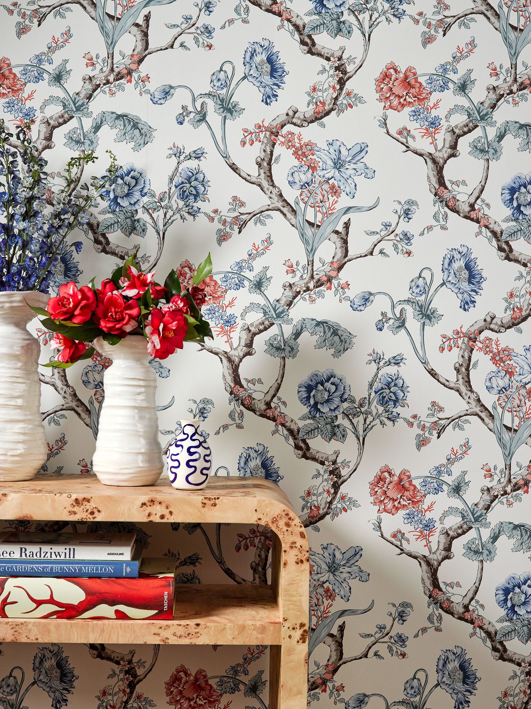 Floral wallpaper with blue, red and brown vines; wooden shelf with ceramic vases and red flowers, plus books below.