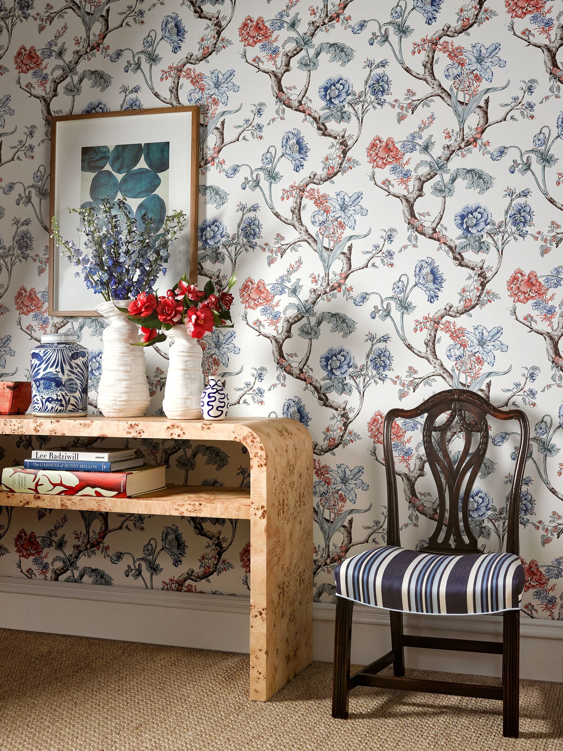 Floral botanical wallpaper in historic noir style; room scene shows a framed print, striped-cushion chair, and blue/white ceramic vases nearby.
