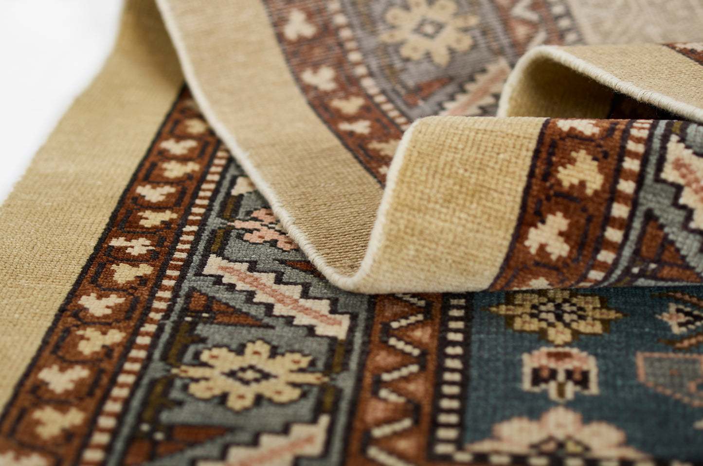Close-up of a traditional rug with beige border, teal and brown geometric motifs, folded to show texture.