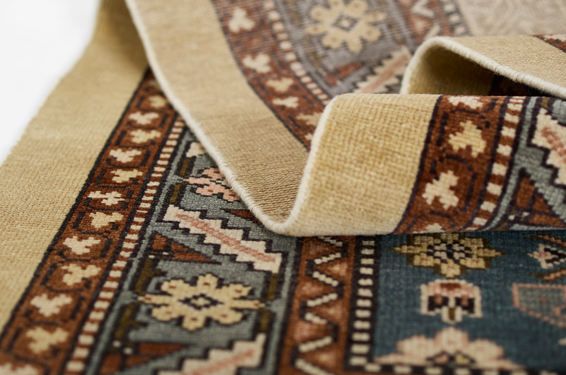 Close-up of a woven rug with beige border, teal field and rust/ivory geometric floral motifs, partially rolled.