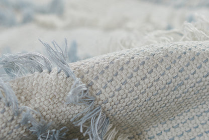 Close-up of a light beige cotton woven throw with pale blue specks and fringe tassels.