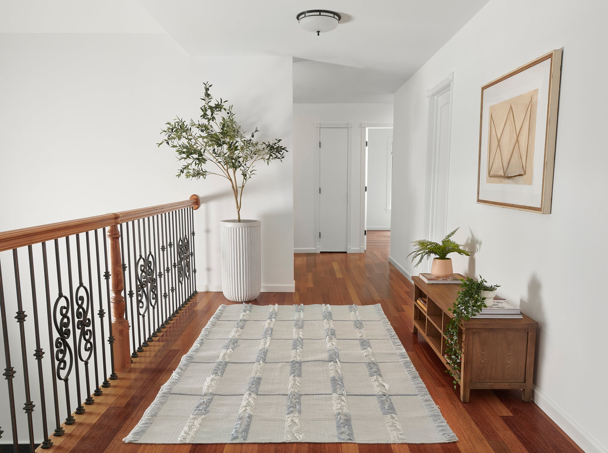 Bright hallway with warm wood floors, black iron railing, tall potted plant, grey striped runner rug, and console with greenery.