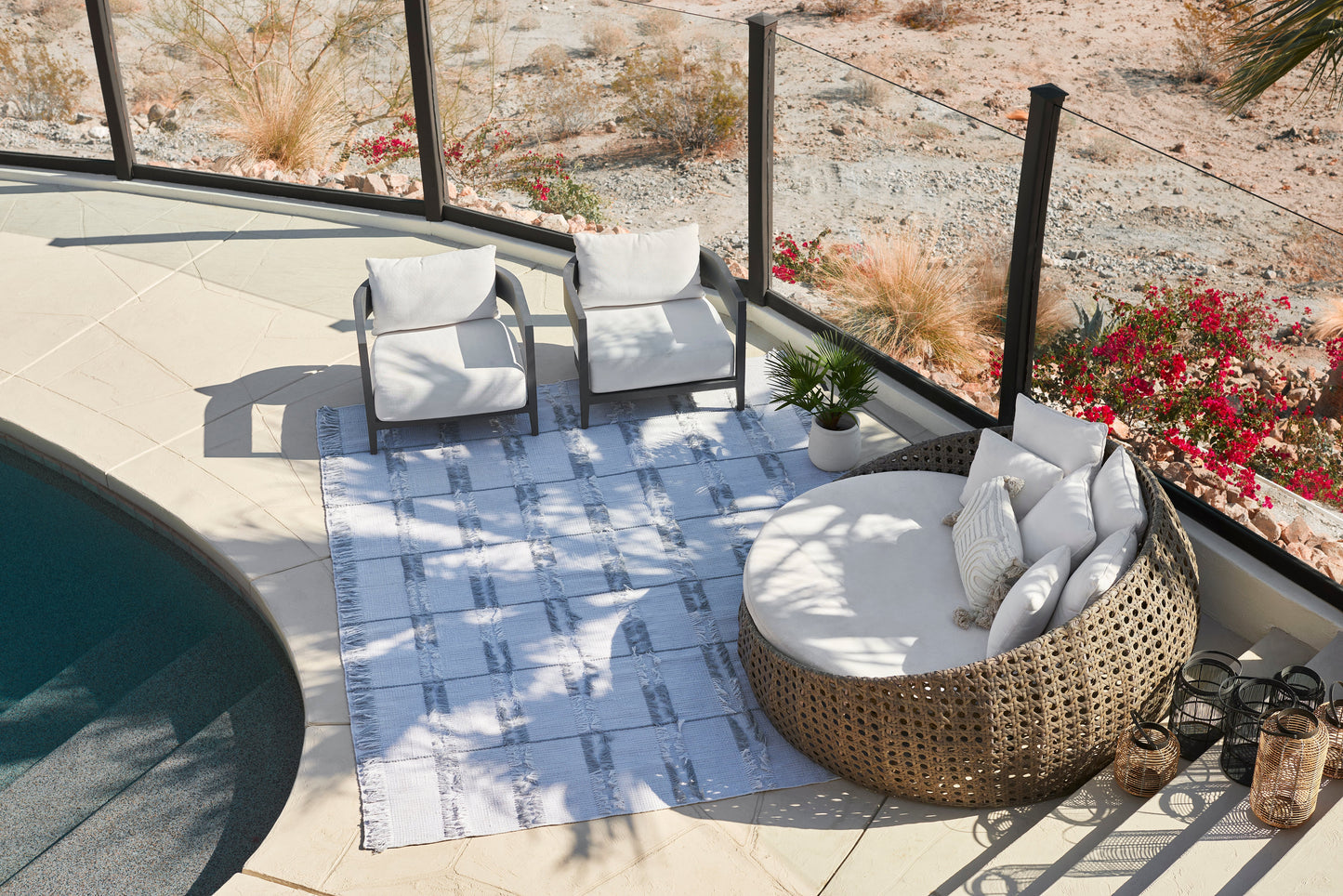 Blue striped 5' x 8' outdoor rug on a poolside patio, with two white-cushion chairs, round wicker lounge, plants and lanterns.