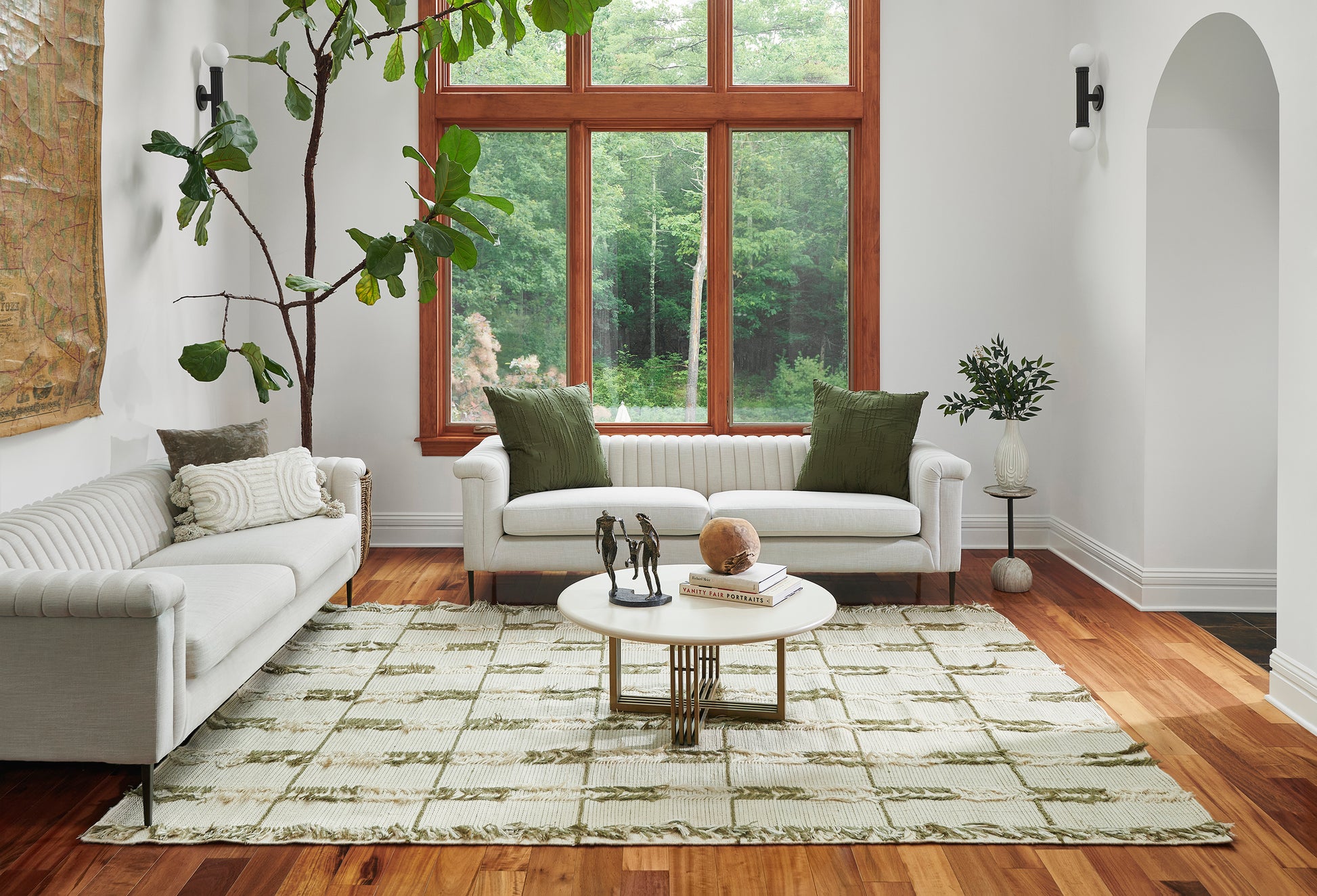 Bright living room with white sofas, olive-green cushions, wooden window frame, green grid rug, round white coffee table.