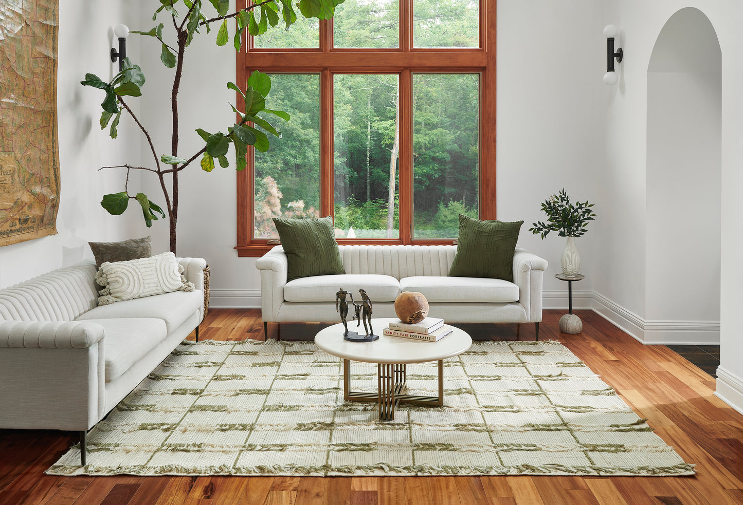 Living room with white sofas & olive-green cushions, wood-framed window, round white coffee table & leafy plants.