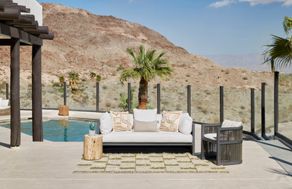 Desert poolside lounge: cream cushions on a dark gray sofa, beige patterned pillows, wood stump table, woven rug, glass railing.