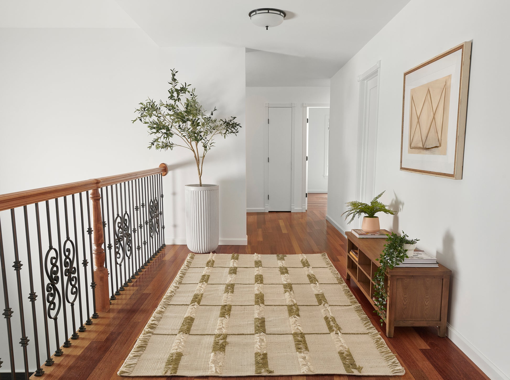 Bright hallway with warm wood floor, beige rug, wooden railing; tall plant, console table with books & framed art.